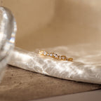 The Verity Diamond Ring with a gold cluster band and sparkling white gemstones rests on a textured, sunlit marble surface, casting delicate patterns of reflected light.