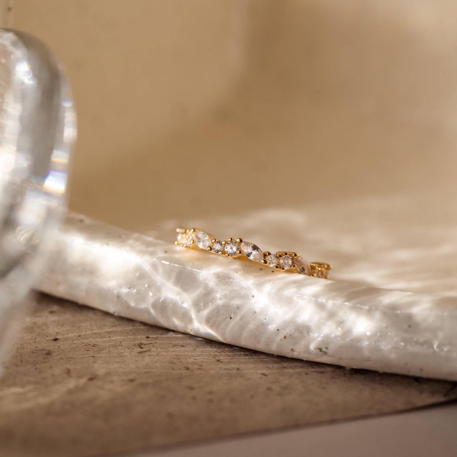 The Verity Diamond Ring with a gold cluster band and sparkling white gemstones rests on a textured, sunlit marble surface, casting delicate patterns of reflected light.