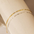 Two Mini Name Bar Bracelets—one gold, one silver—each with an engraved name on a rectangular plate in a modern paperclip chain style. A thoughtful, personalized gift.