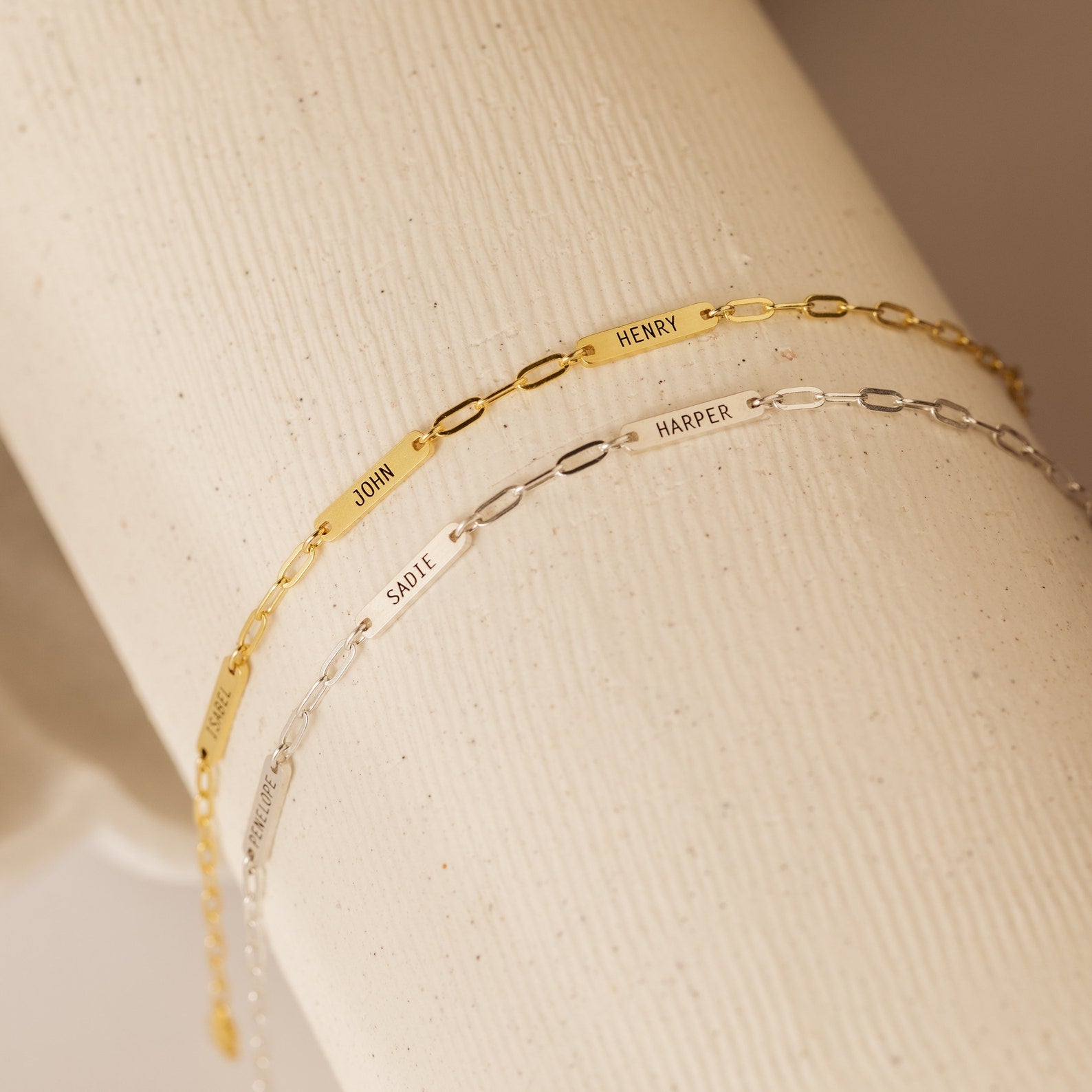 Two Mini Name Bar Bracelets—one gold, one silver—each with an engraved name on a rectangular plate in a modern paperclip chain style. A thoughtful, personalized gift.