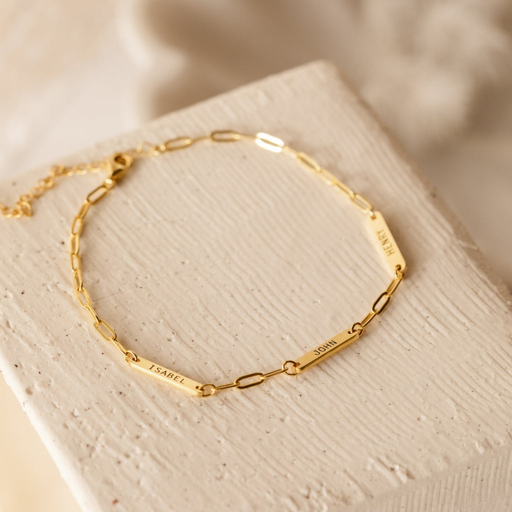 The Mini Name Bar Bracelet is a gold paperclip bracelet with engraved bars reading ISABEL, JOHN, and HENRY on a textured surface—an elegant personalized accessory, perfect as a customized gift.