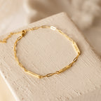 The Mini Name Bar Bracelet is a gold paperclip bracelet with engraved bars reading ISABEL, JOHN, and HENRY on a textured surface—an elegant personalized accessory, perfect as a customized gift.