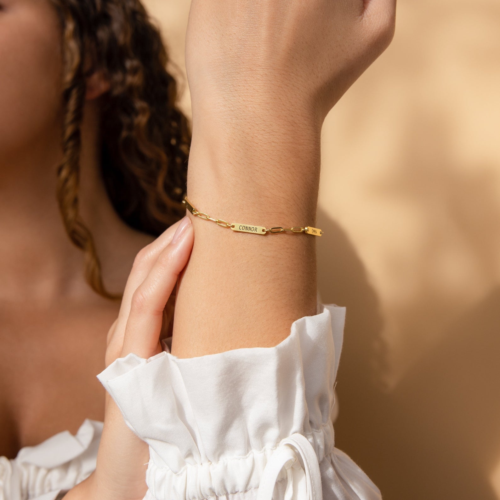 A person wearing a white blouse showcases the Mini Name Bar Bracelet on their wrist against a beige background.