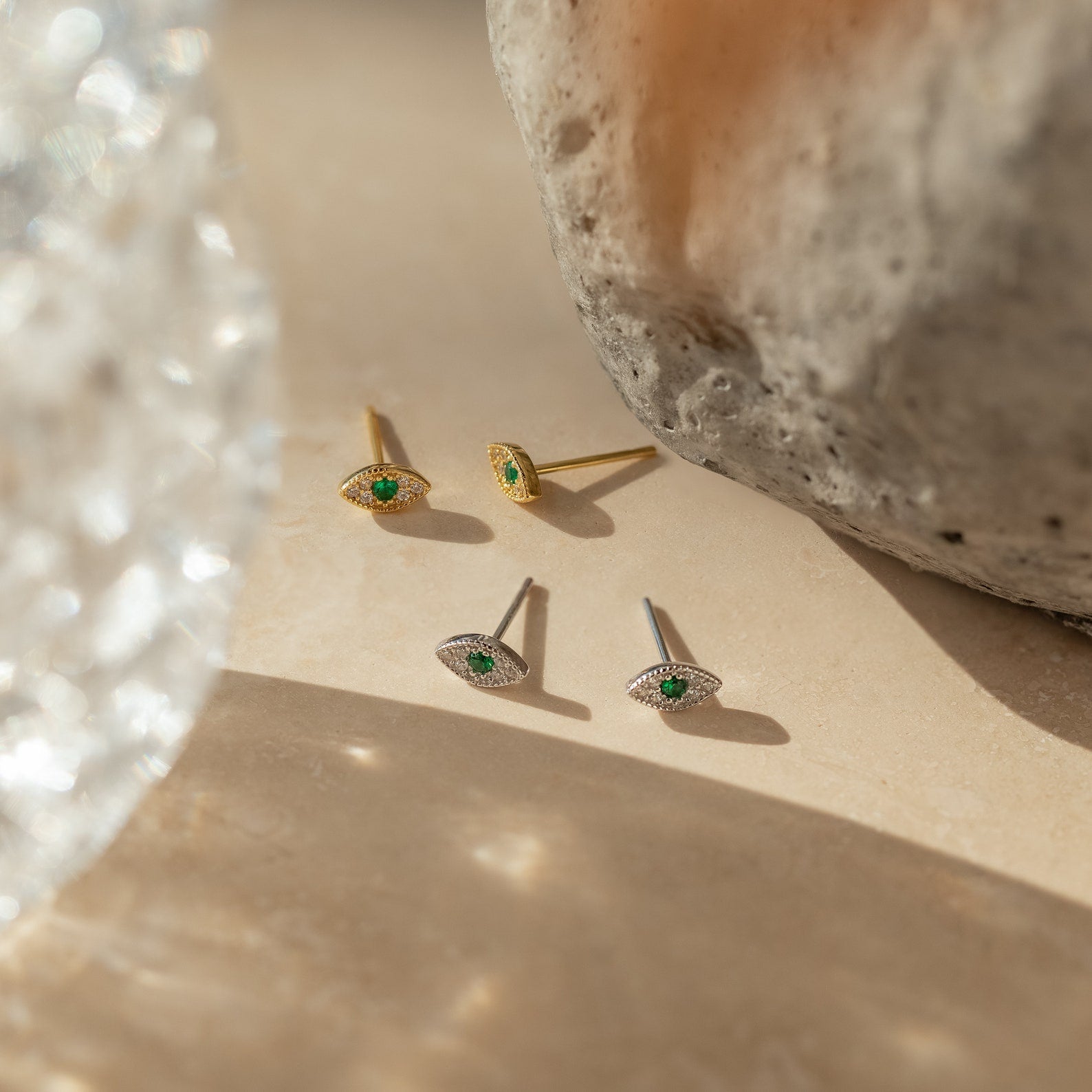 Four Haven Emerald Eye Studs with emerald stones and gold accents, featuring an Evil Eye design, are displayed on a beige surface alongside rocks.