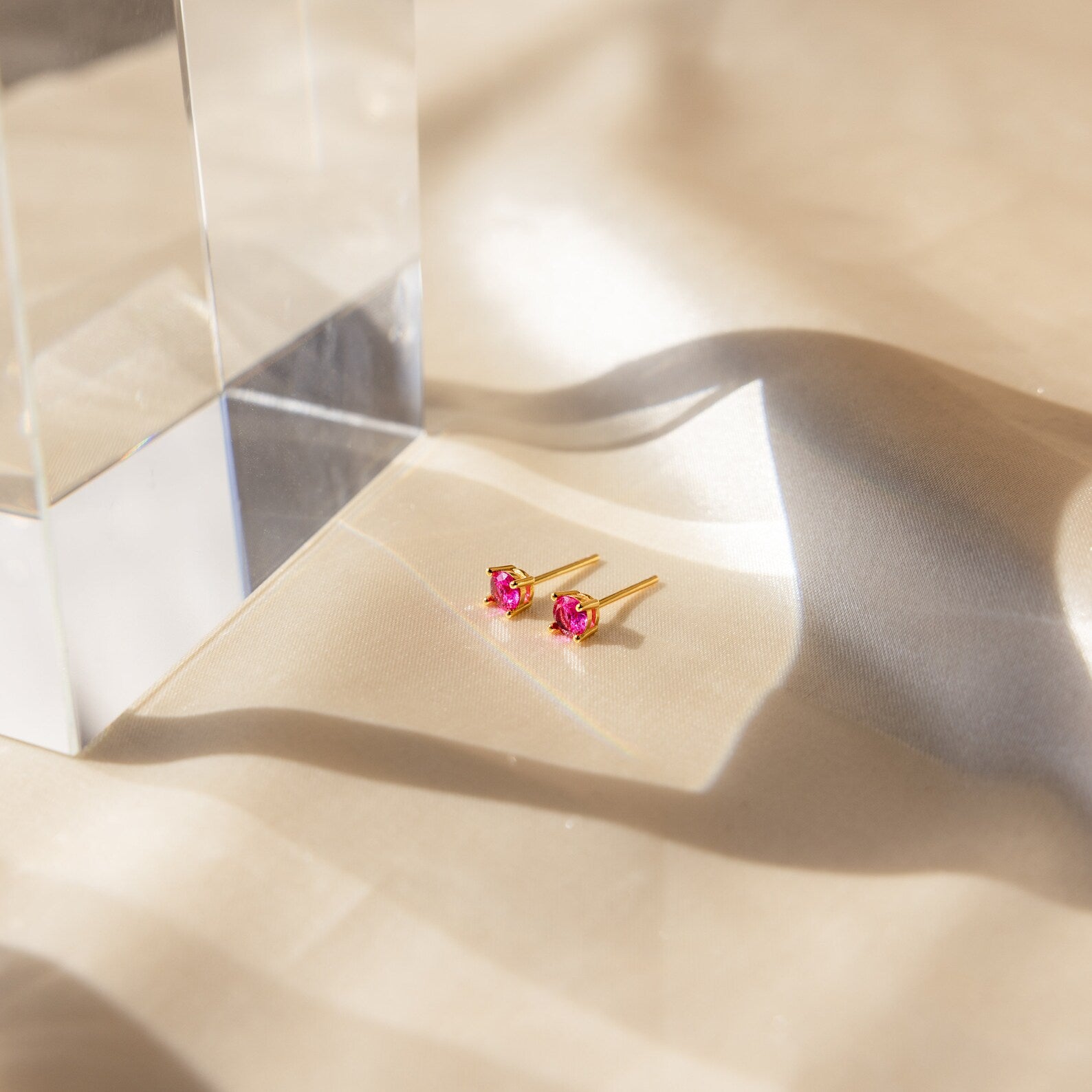 Booker Large Birthstone Studs with pink gemstones rest on beige satin, softly illuminated by sunlight and shadows—these earrings are a thoughtful jewelry gift.