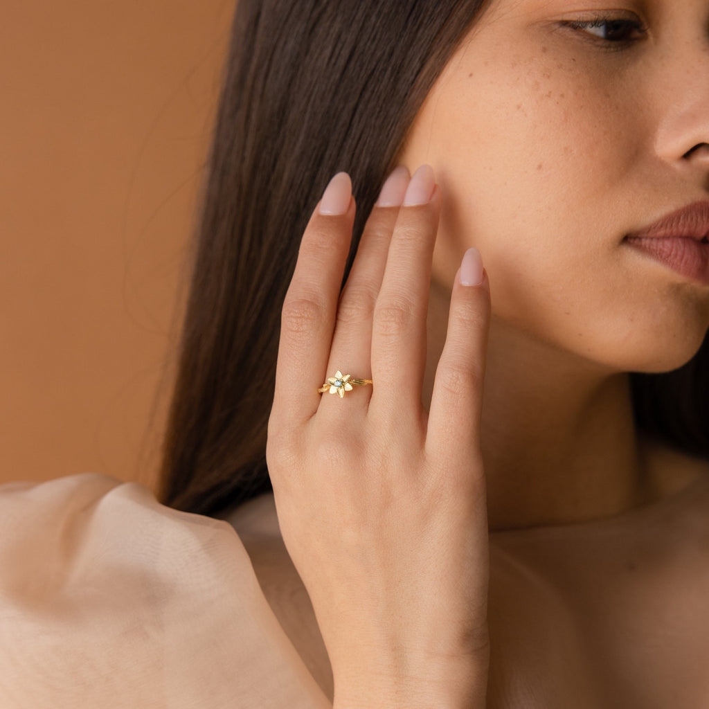A woman with long brown hair touches her face, wearing the Flower Birthstone Ring featuring a vintage-inspired gold flower design and a sheer beige top.