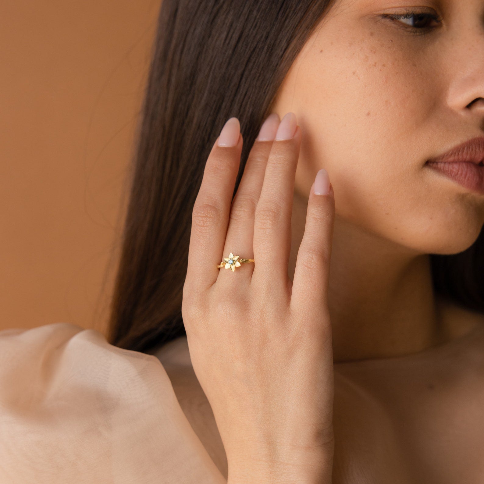 A woman with long brown hair touches her face, wearing the Flower Birthstone Ring featuring a vintage-inspired gold flower design and a sheer beige top.
