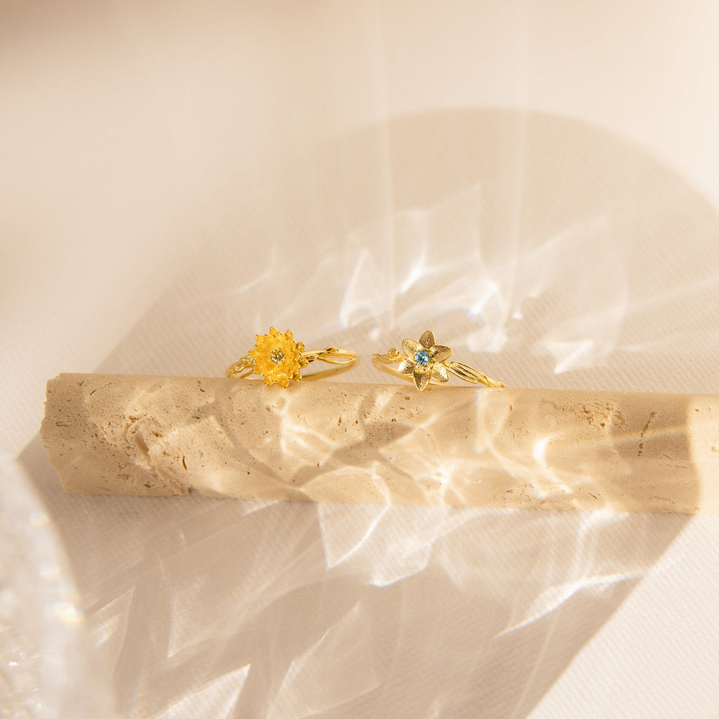 A Flower Birthstone Ring with a vintage-inspired flower design rests on a textured beige surface in soft sunlight.