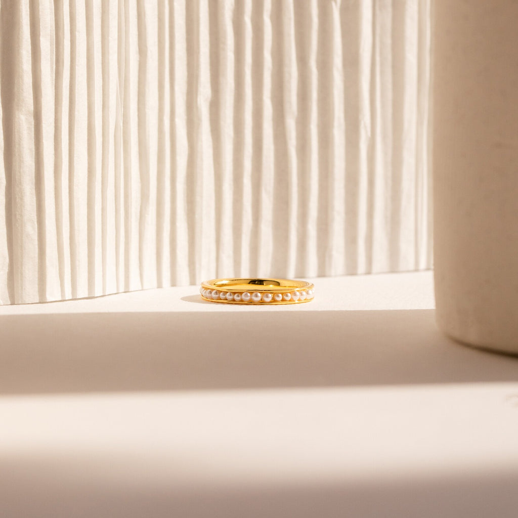 The Alicia Pearl Inlay Band, a minimalist ring featuring delicate pearls, is showcased on a neutral surface with soft natural light highlighting its elegant design against a textured backdrop.