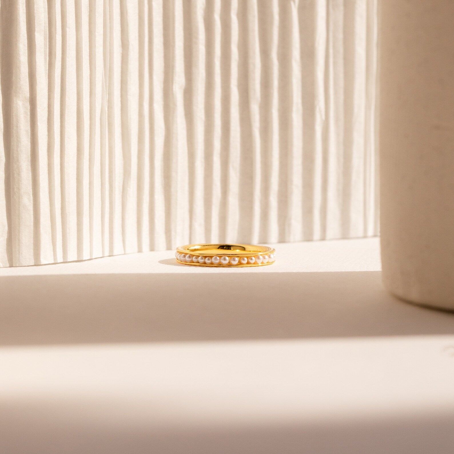 The Alicia Pearl Inlay Band, a minimalist ring featuring delicate pearls, is showcased on a neutral surface with soft natural light highlighting its elegant design against a textured backdrop.