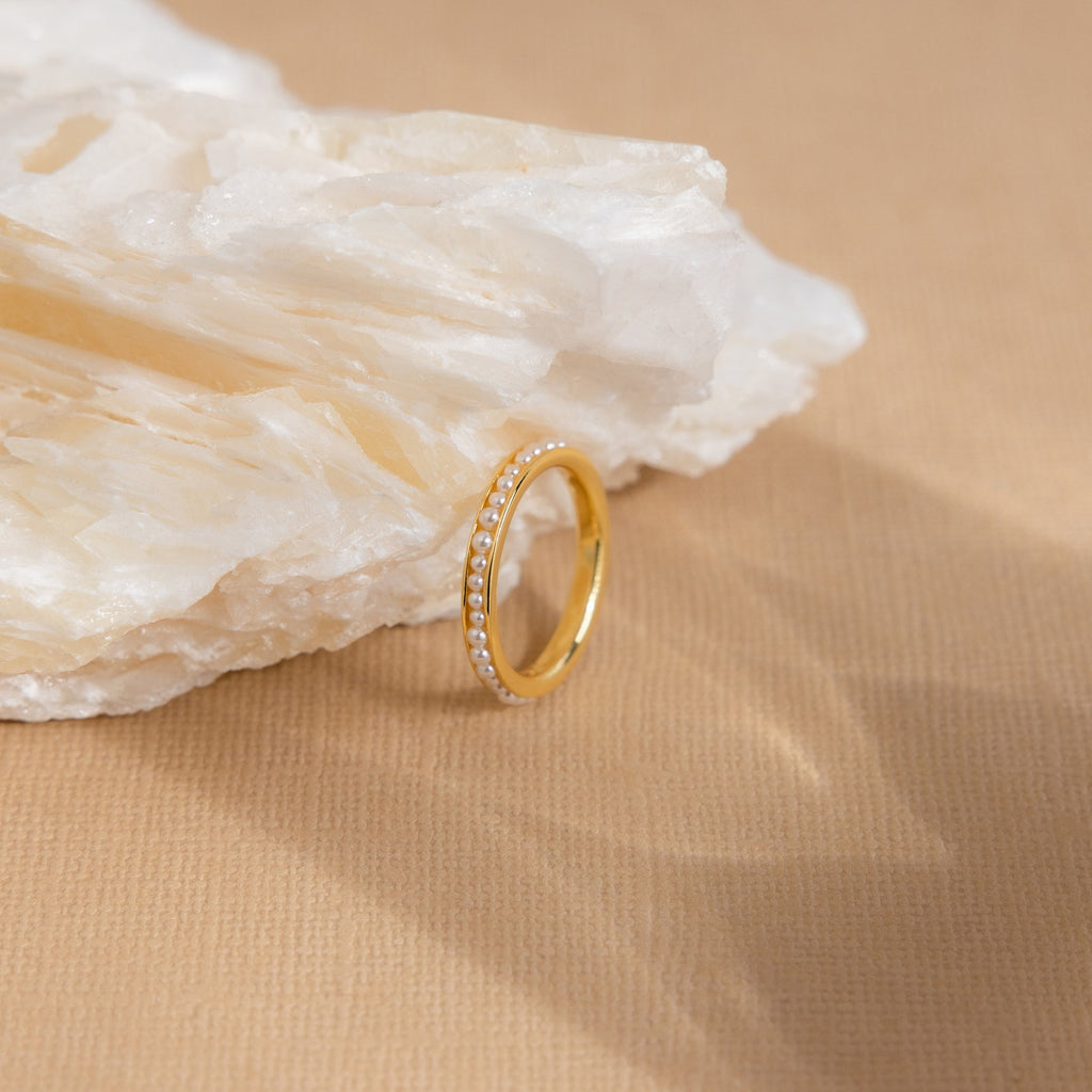 The Alicia Pearl Inlay Band, a minimalist gold ring with pearl accents, rests against a white stone on a beige textured surface.