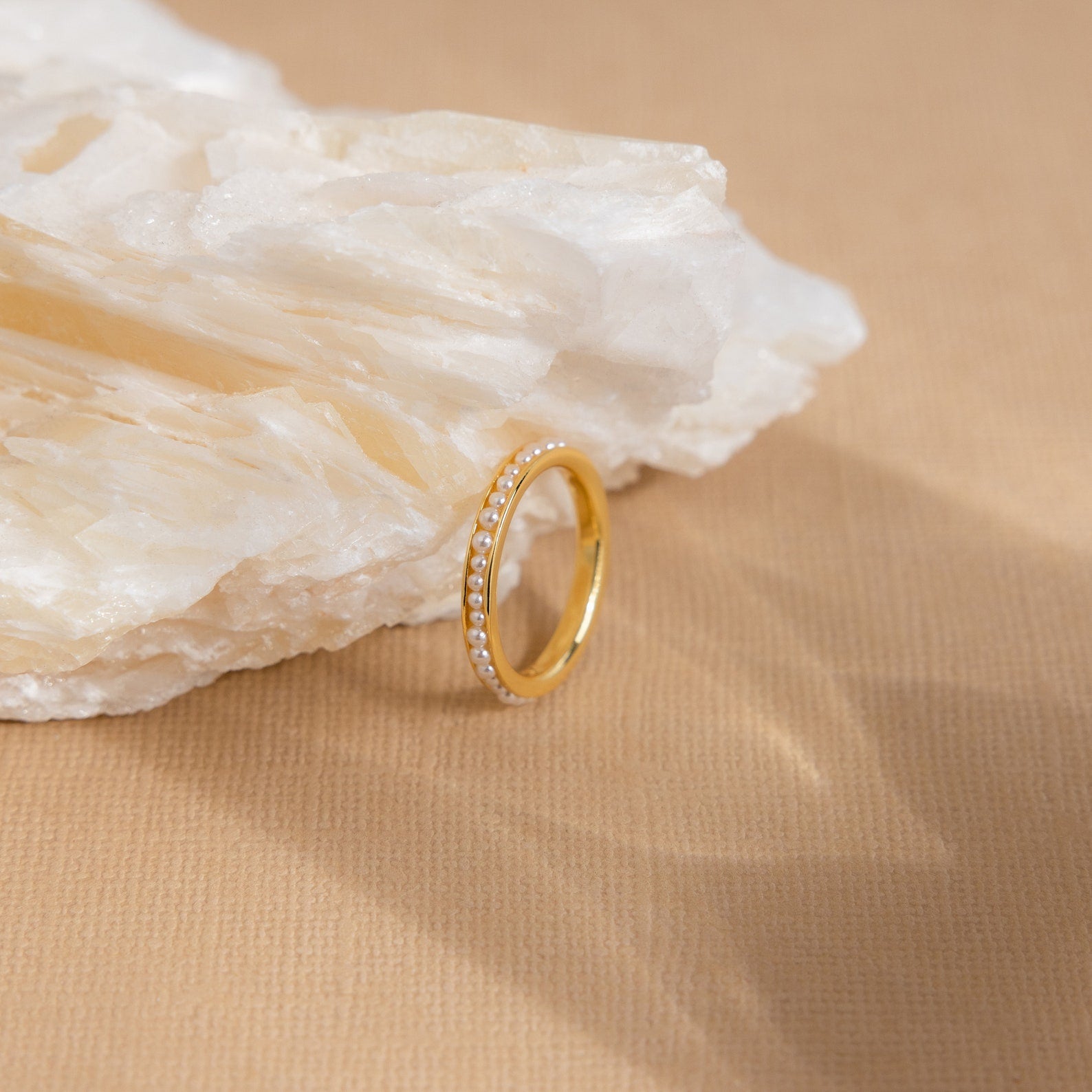 The Alicia Pearl Inlay Band, a minimalist gold ring with pearl accents, rests against a white stone on a beige textured surface.