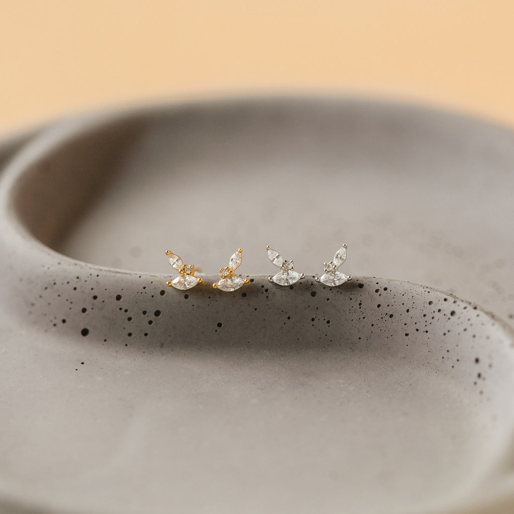 Two pairs of Flora Marquise Studs, one in gold and one in silver, are displayed on a speckled ceramic dish—perfect for fans of minimalist, leaf-inspired earrings.