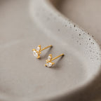 The Flora Marquise Studs in 18K Gold, featuring three clear gemstones, are displayed on a smooth ceramic dish—simple yet elegant wedding jewelry for any occasion.