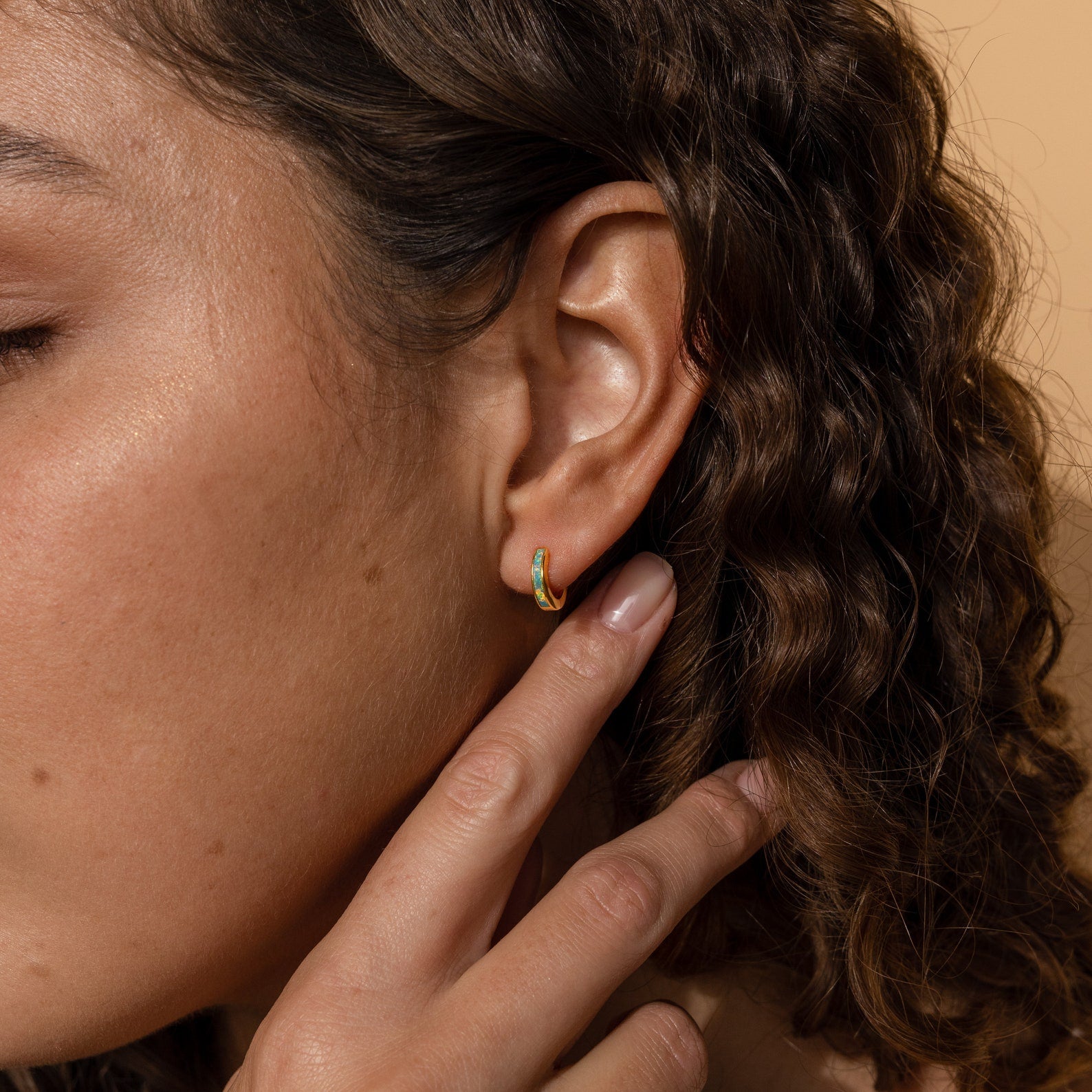 A close-up of a woman's ear wearing the Opal Inlay Huggies Set, showcasing the hoops' vibrant opal colors.