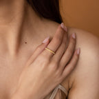 A woman's hand with nude nails rests on her shoulder, showcasing the Alicia Pearl Inlay Band, a delicate gold stackable ring adorned with small gemstones.