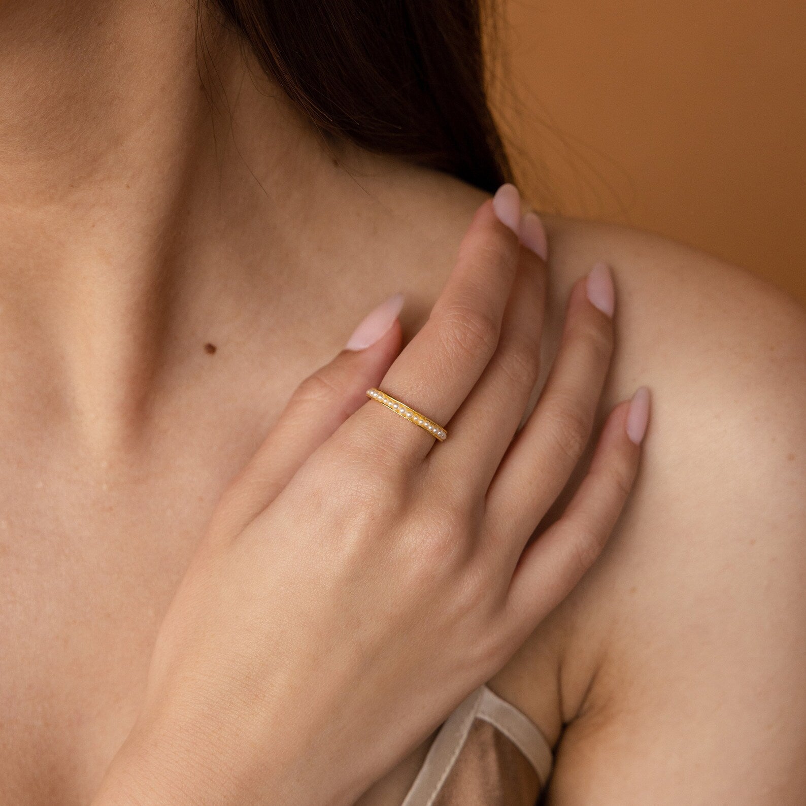 A woman's hand with nude nails rests on her shoulder, showcasing the Alicia Pearl Inlay Band, a delicate gold stackable ring adorned with small gemstones.