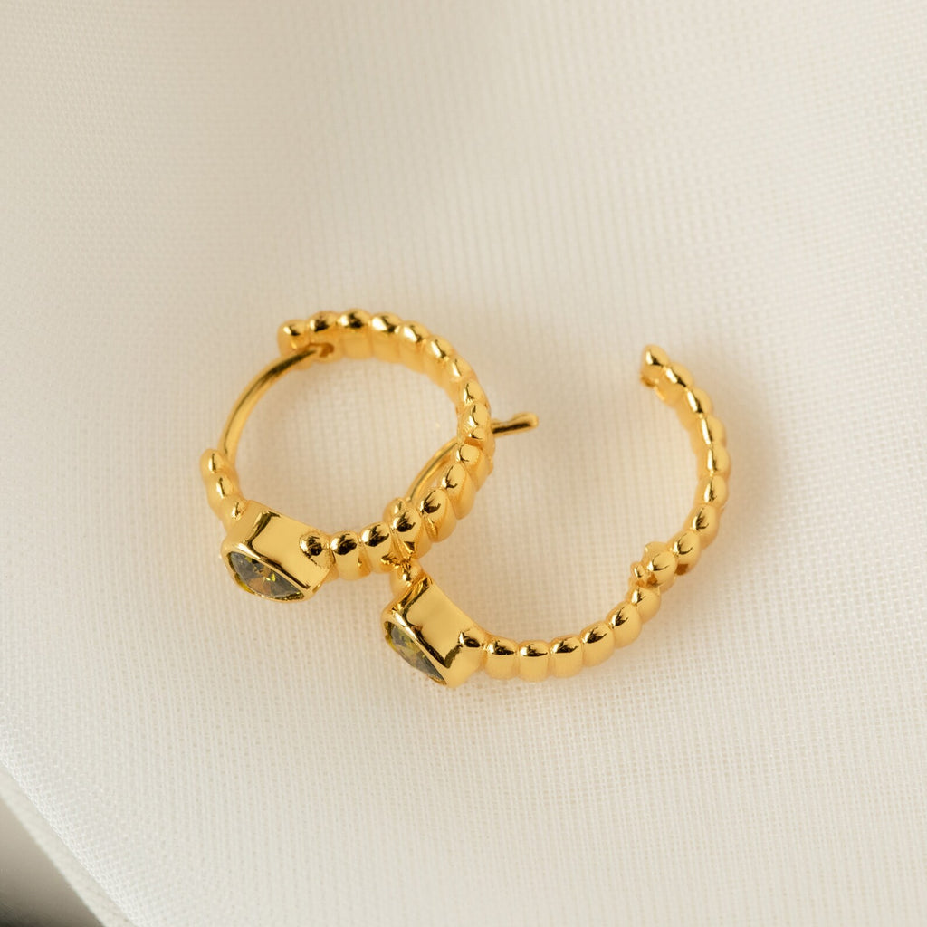 Peridot Heart Huggies—gold hoop earrings with heart-shaped peridot stones, shown on a cream fabric background. Perfect minimalistic jewelry to elevate your style.