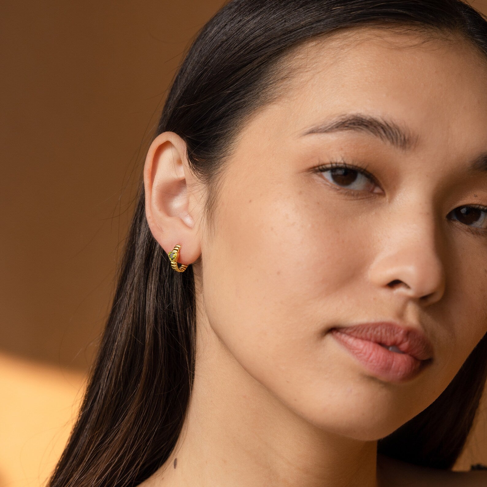 Woman with long dark hair wears Peridot Heart Huggies earrings, gazing slightly to the side against a brown background.