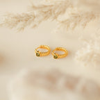 A pair of gold rings with a green gem, inspired by the minimalistic Peridot Heart Huggies earrings.