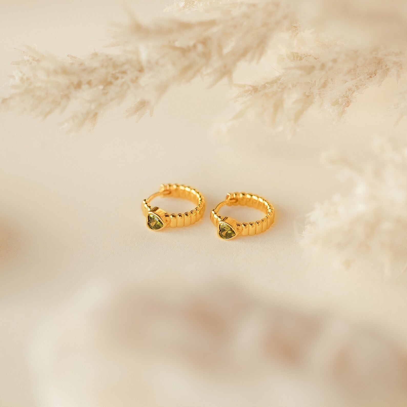 A pair of gold rings with a green gem, inspired by the minimalistic Peridot Heart Huggies earrings.