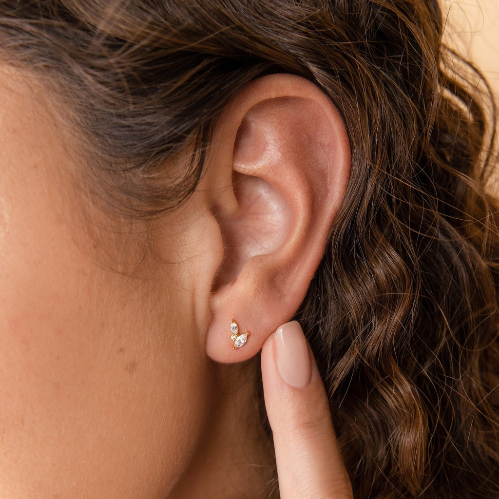 A hand touches a woman's ear adorned with Flora Marquise Studs in 18K Gold, minimalist earrings featuring tiny stones set in a delicate gold design.