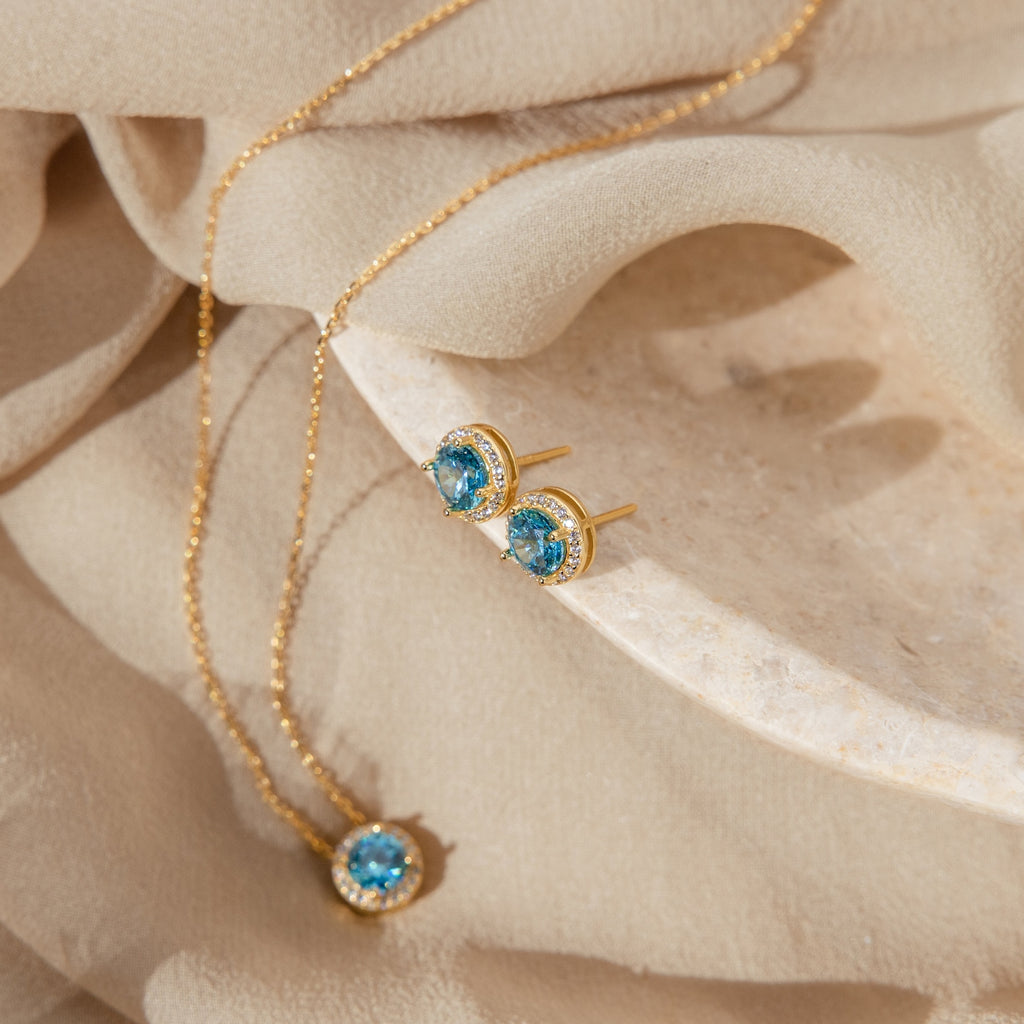 Halo Birthstone Studs with blue gemstones are displayed on beige fabric and a stone dish, paired with vintage-inspired earrings and a gold necklace for an elegant accent.