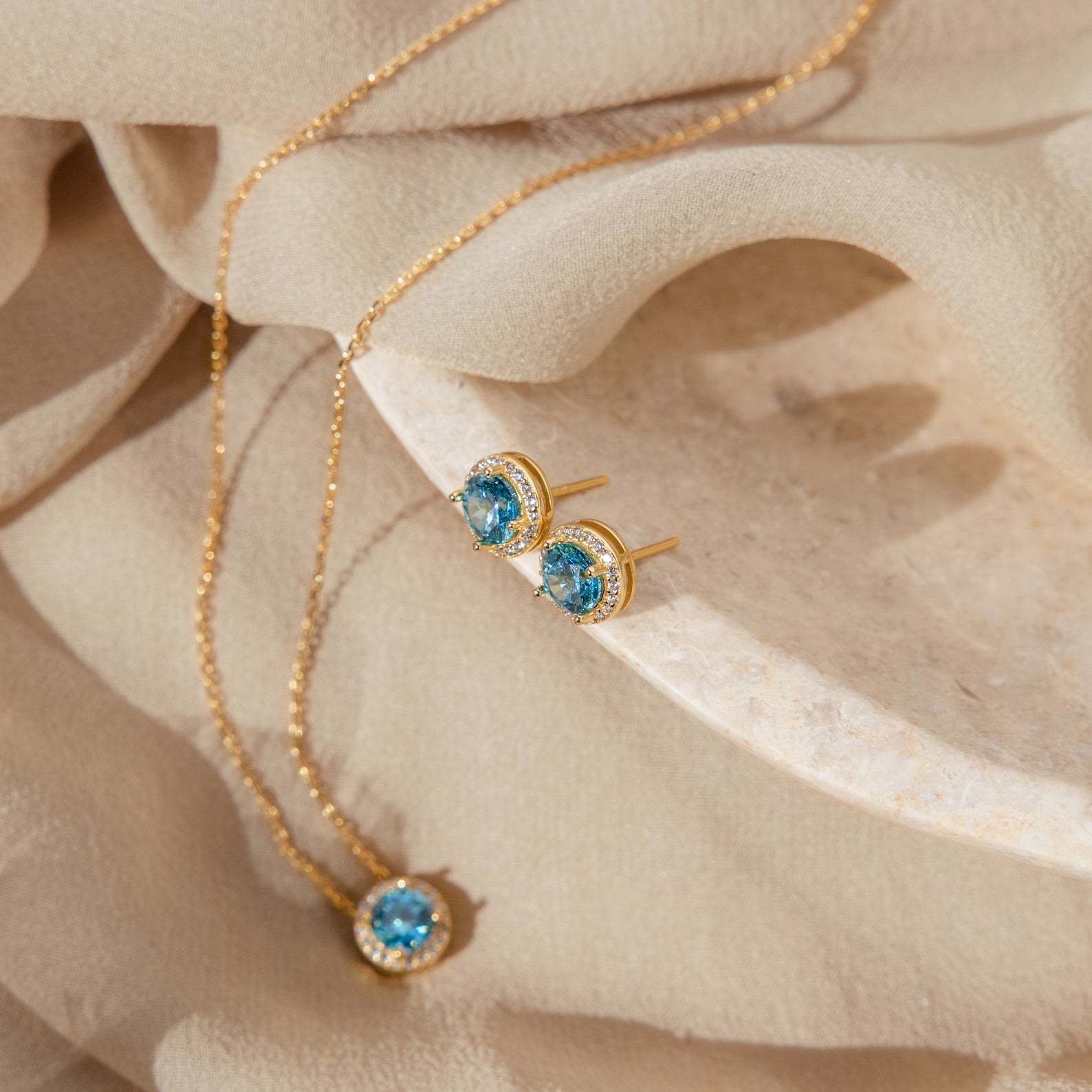 Halo Birthstone Studs with blue gemstones are displayed on beige fabric and a stone dish, paired with vintage-inspired earrings and a gold necklace for an elegant accent.