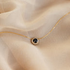 The Halo Birthstone Necklace, featuring a black gemstone pendant and statement design, is showcased on light beige fabric.