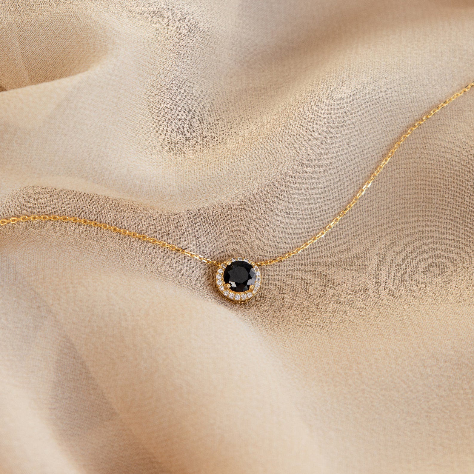 The Halo Birthstone Necklace, featuring a black gemstone pendant and statement design, is showcased on light beige fabric.