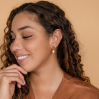 Woman with curly hair smiles in an orange top and earthy jewelry—Carnelian Pearl Earrings featuring orange and pearl dangles—posed against a beige background.