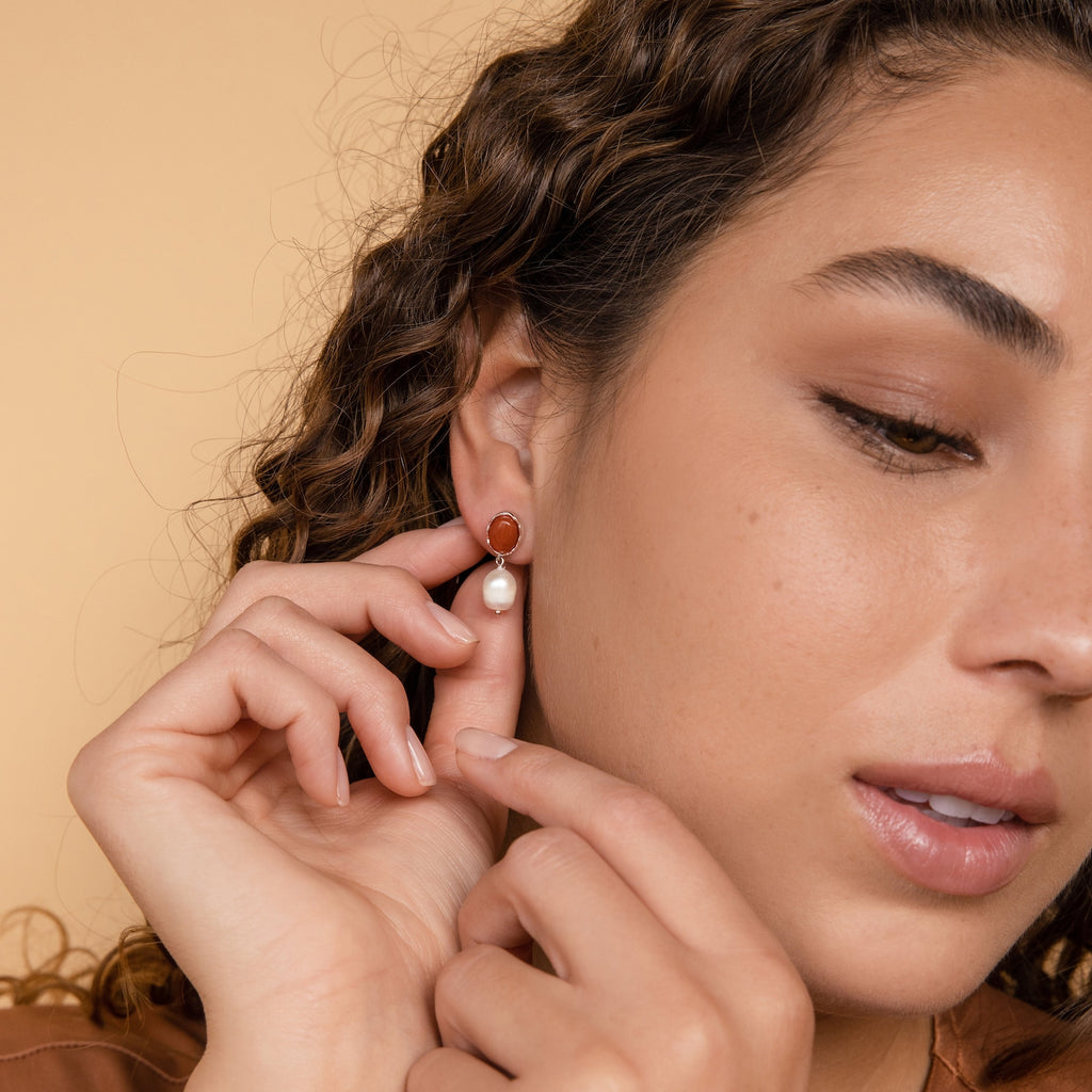 A woman with curly hair puts on Carnelian Pearl Earrings, featuring a pearl dangle and vibrant red stone, showcasing elegant earthy jewelry against a beige background.