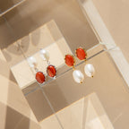 Two pairs of Carnelian Pearl Earrings, featuring oval red carnelian stones and white pearls, rest on a clear geometric surface—ideal for those who love earthy jewelry.