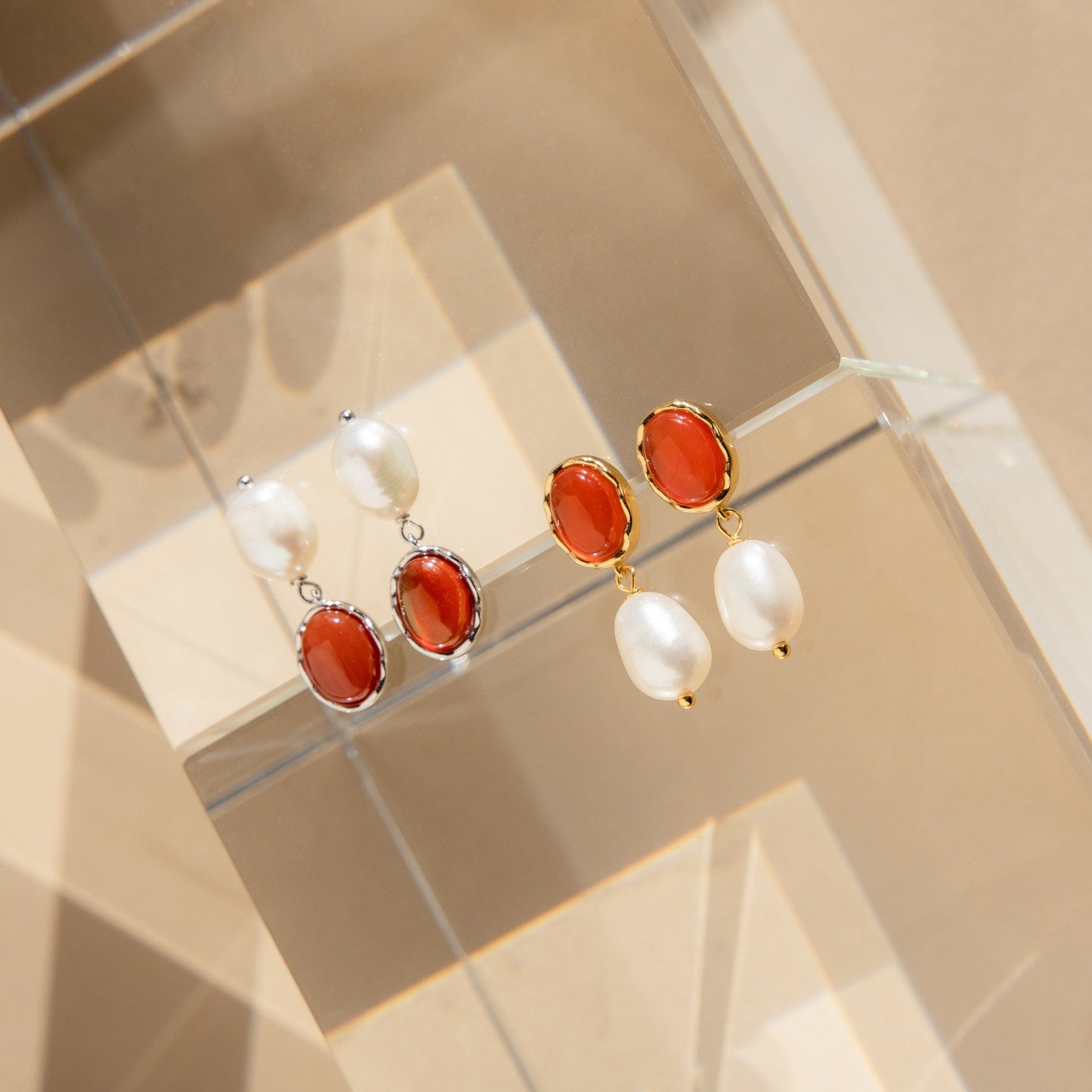 Two pairs of Carnelian Pearl Earrings, featuring oval red carnelian stones and white pearls, rest on a clear geometric surface—ideal for those who love earthy jewelry.