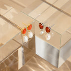 Carnelian Pearl Earrings are showcased on clear glass stands, featuring vibrant orange carnelian stones and pearl dangles for an elegant, earthy touch.