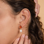 A close-up of an ear wearing Carnelian Pearl Earrings, featuring a gold setting with an orange carnelian stone and a dangling white pearl, capturing the elegance of pearl dangle earrings and earthy jewelry style.