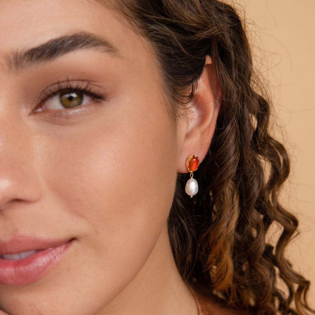 A woman with curly hair wears Carnelian Pearl Earrings—red and gold with a dangling white pearl—an elegant, earthy jewelry style showcased in a close-up of her face.