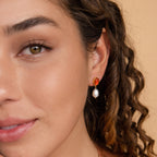 A woman with curly hair wears Carnelian Pearl Earrings—red and gold with a dangling white pearl—an elegant, earthy jewelry style showcased in a close-up of her face.