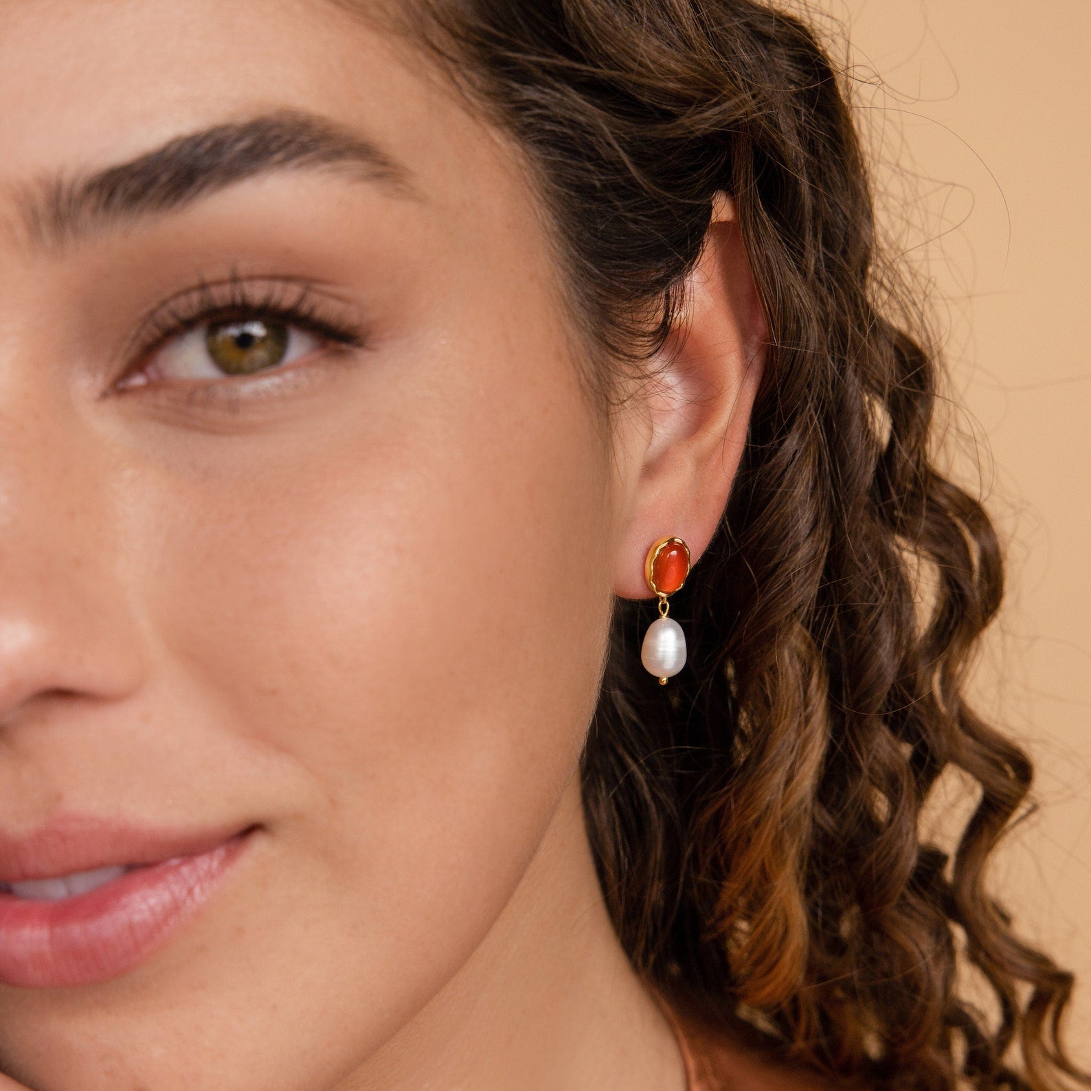 A woman with curly hair wears Carnelian Pearl Earrings—red and gold with a dangling white pearl—an elegant, earthy jewelry style showcased in a close-up of her face.