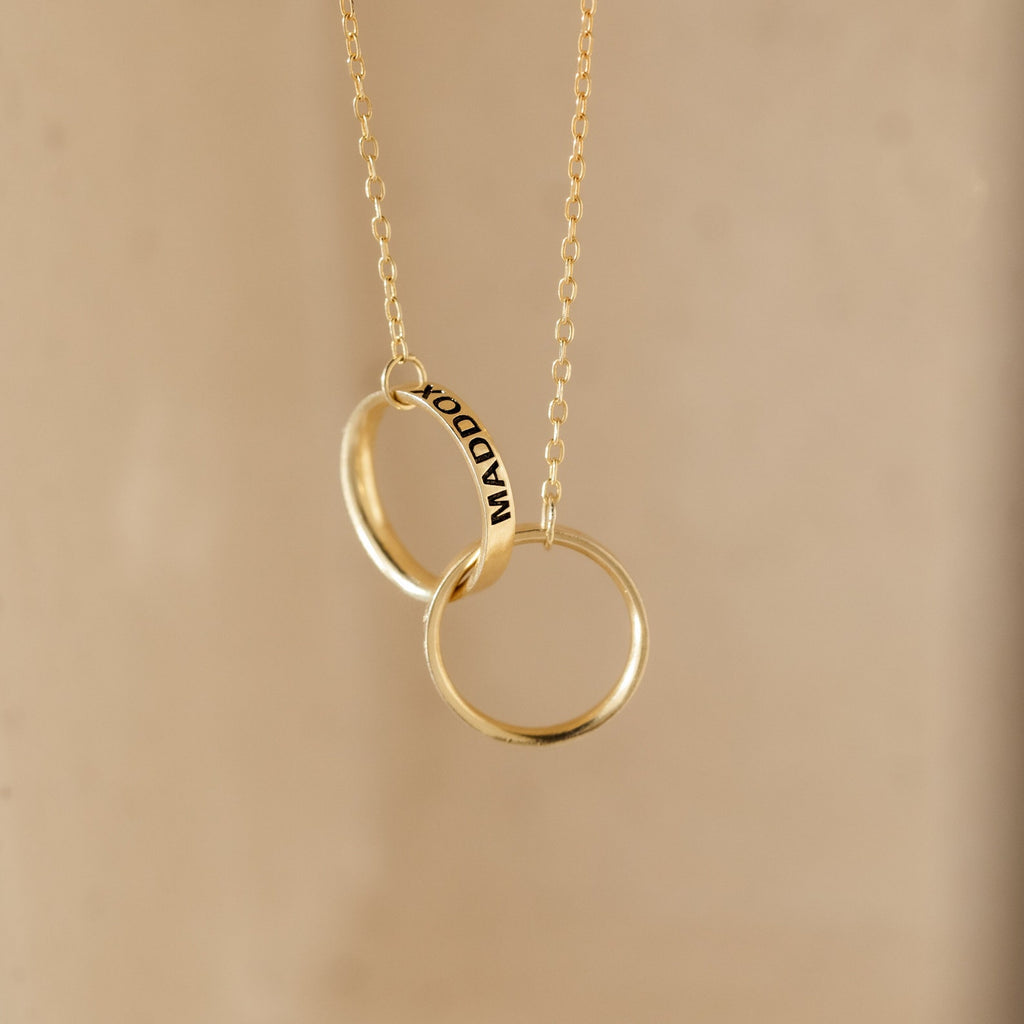 The Engraved Interlocking Necklace features two gold rings, one personalized with MADDOX, displayed on a neutral background. A timeless anniversary gift designed to be cherished forever.