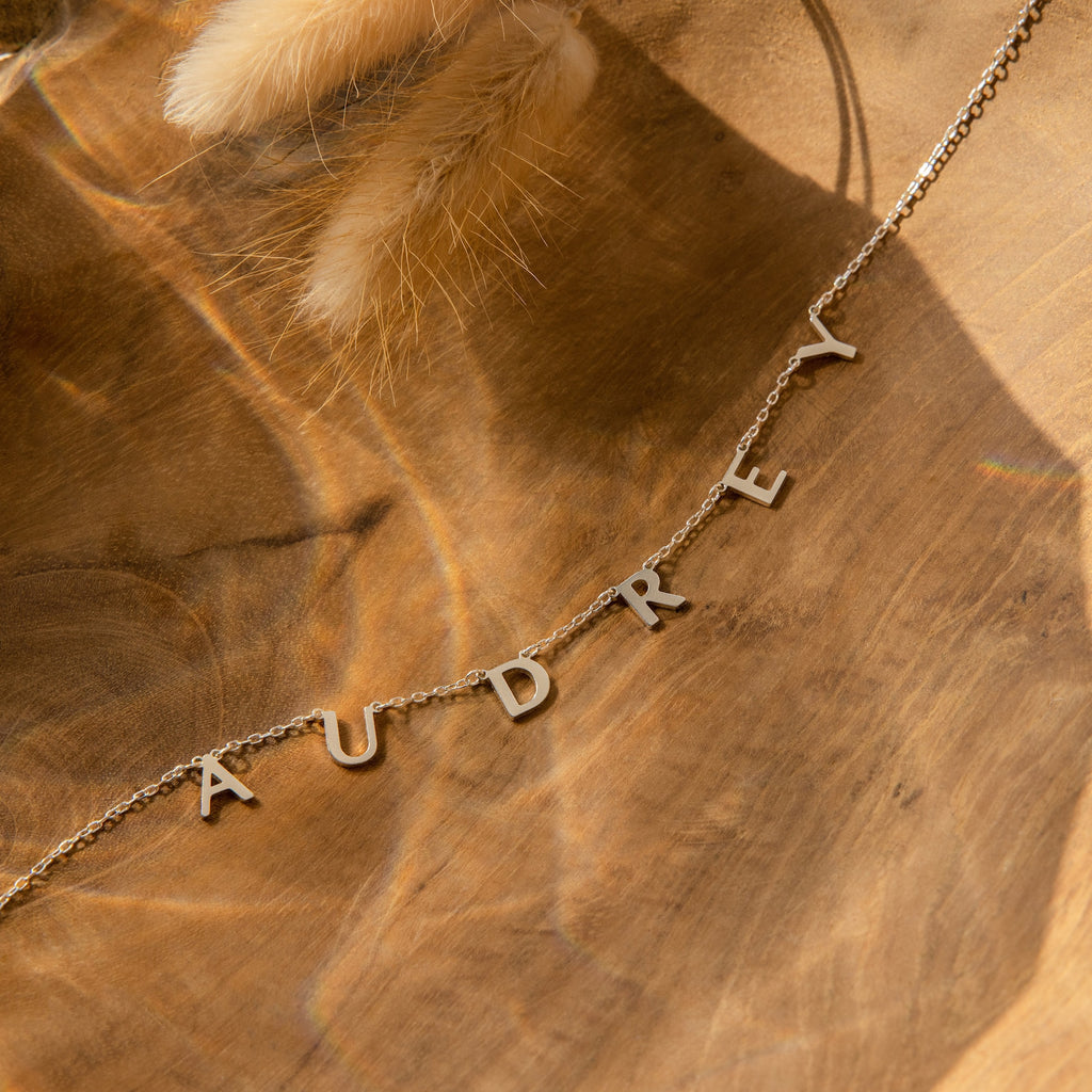 The Apollo Letter Name Necklace features gold letter charms spelling AUDREY, shown on a wooden background with soft lighting and dried grass—an ideal custom initial necklace for adding a unique, personalized touch.