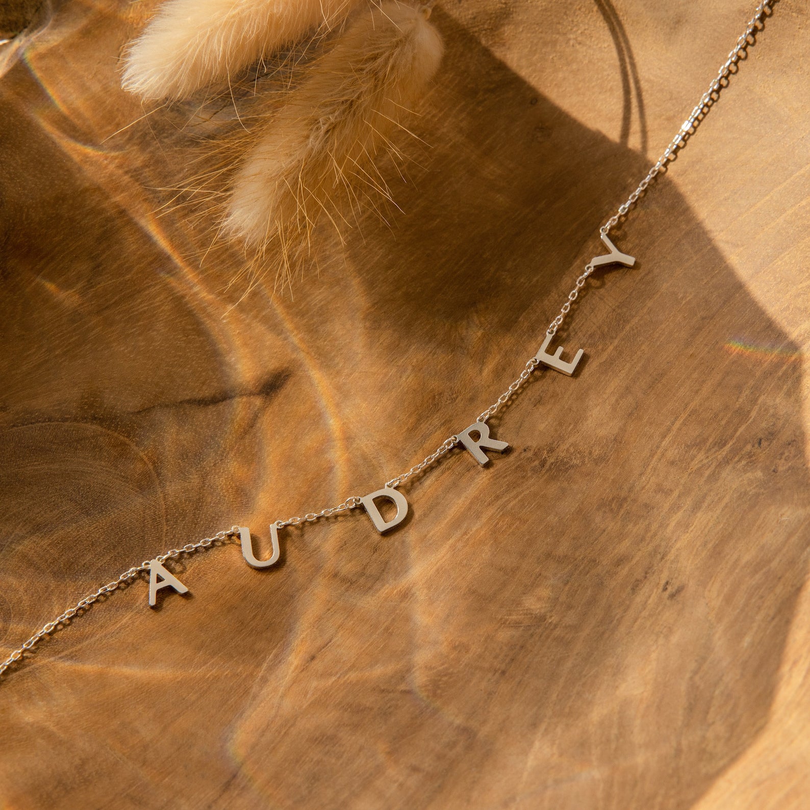 The Apollo Letter Name Necklace features gold letter charms spelling AUDREY, shown on a wooden background with soft lighting and dried grass—an ideal custom initial necklace for adding a unique, personalized touch.