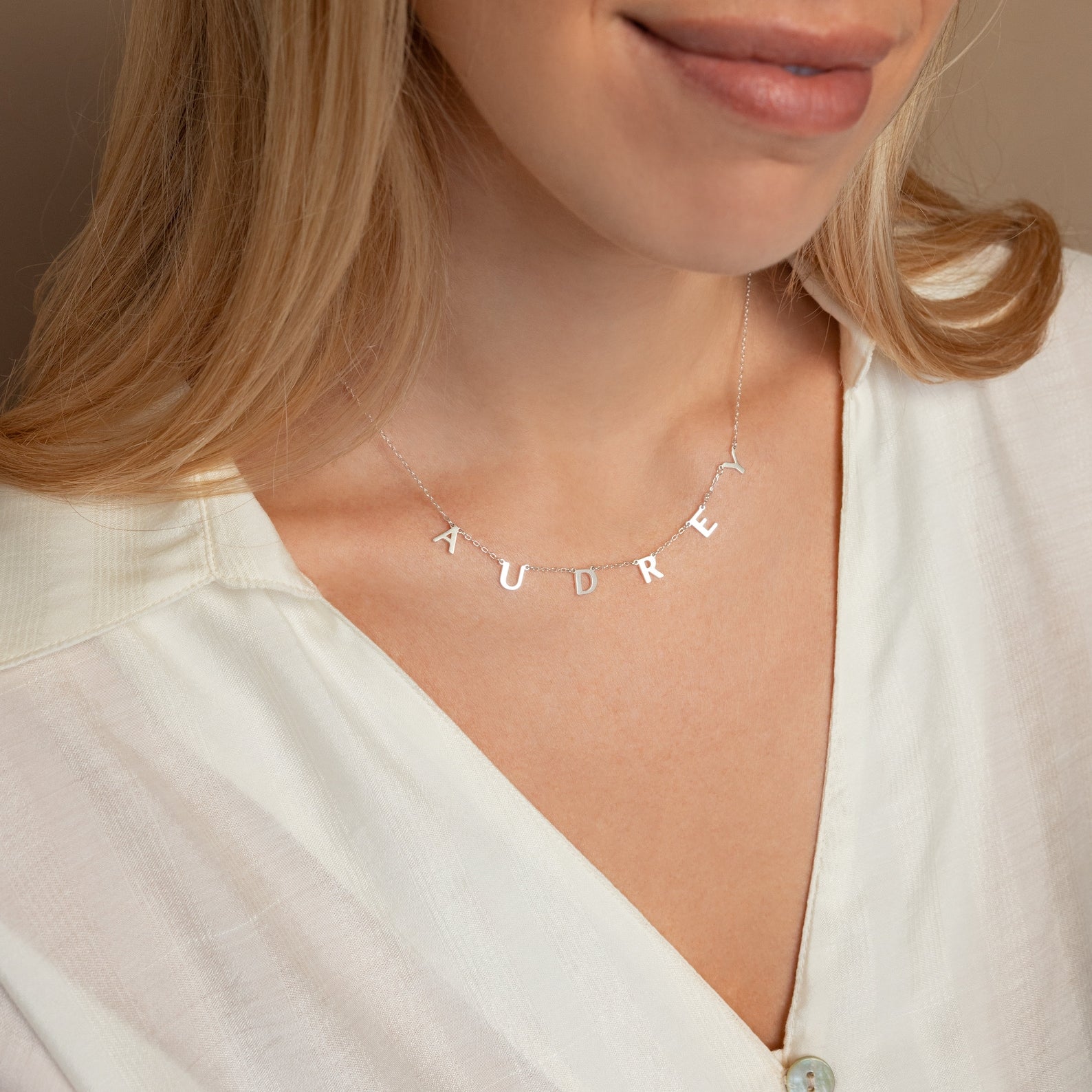 A woman wears a white blouse and the Apollo Letter Name Necklace in silver, personalized with "AUDREY," highlighting its chic, minimalist style.