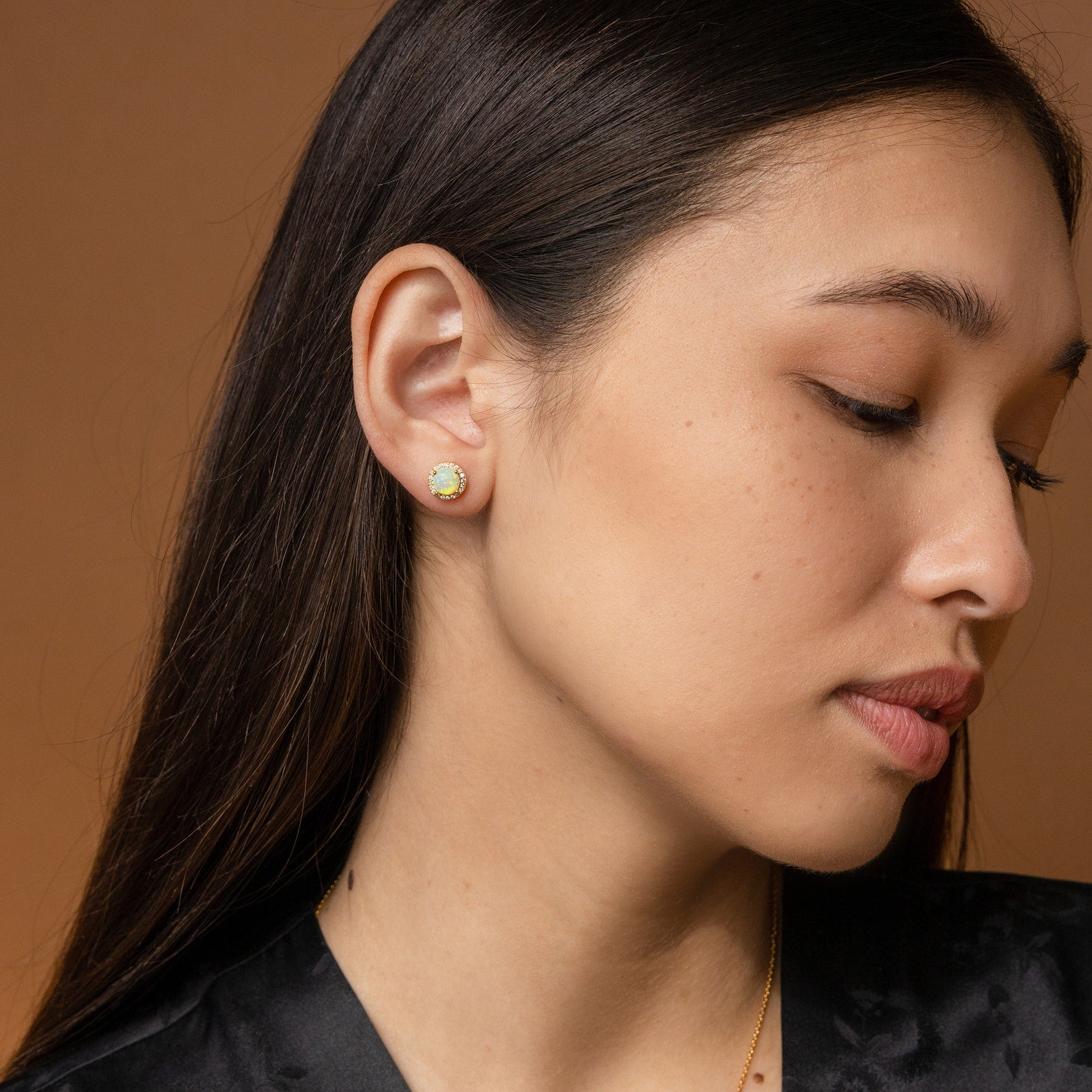 A woman with long dark hair wears Halo Birthstone Studs, gazing down against a brown backdrop.