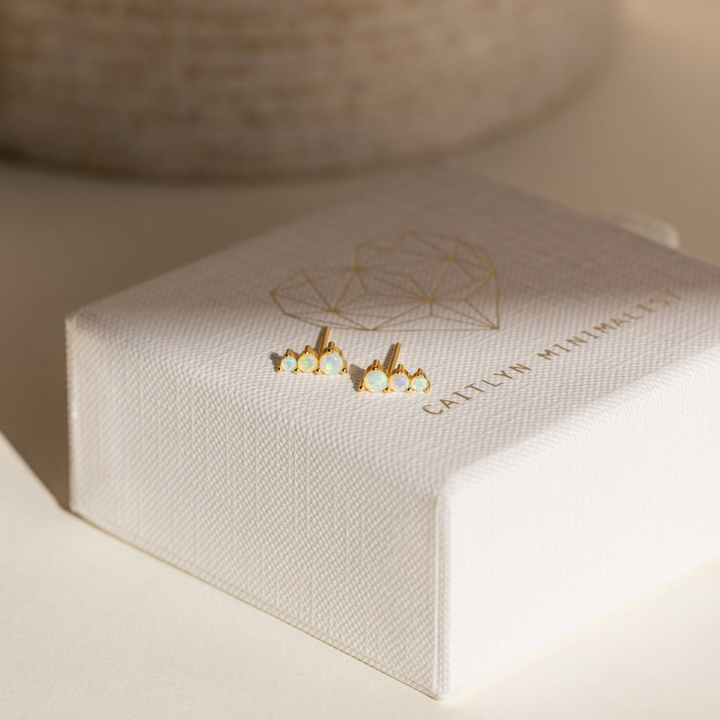 Triple Birthstone Studs adorned with small white stones, these gold stud earrings are a meaningful gift for mothers and daughters, displayed on a textured white jewelry box with a gold design—perfect as birthstone earrings.