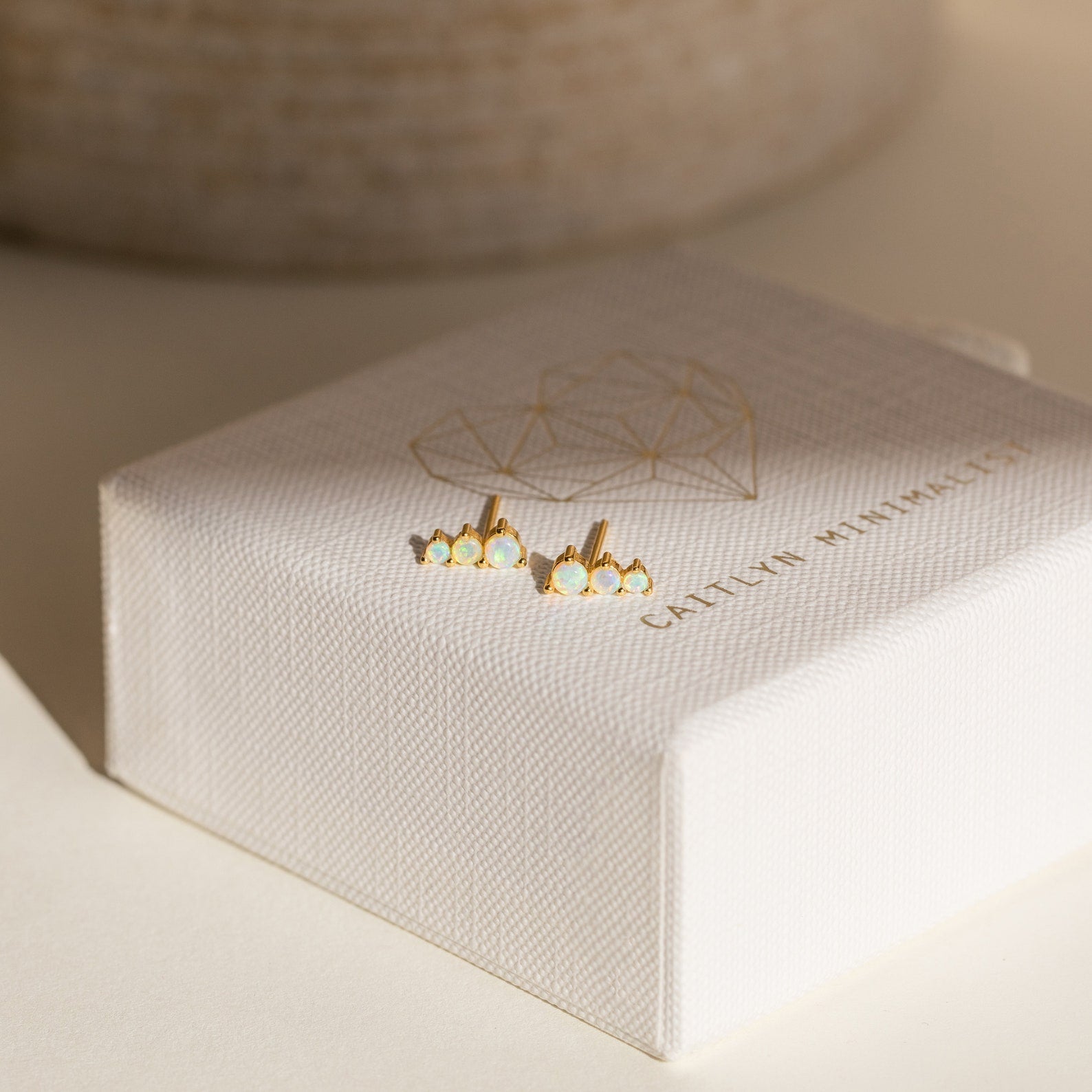 Triple Birthstone Studs adorned with small white stones, these gold stud earrings are a meaningful gift for mothers and daughters, displayed on a textured white jewelry box with a gold design—perfect as birthstone earrings.