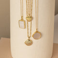 Four gold necklaces with differently shaped pendants, including the Pearl Disc Locket Necklace, are elegantly displayed on a textured cylindrical off-white surface—making them perfect for meaningful gifting.