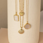 Four gold necklaces with differently shaped pendants, including the Pearl Disc Locket Necklace, are elegantly displayed on a textured cylindrical off-white surface—making them perfect for meaningful gifting.