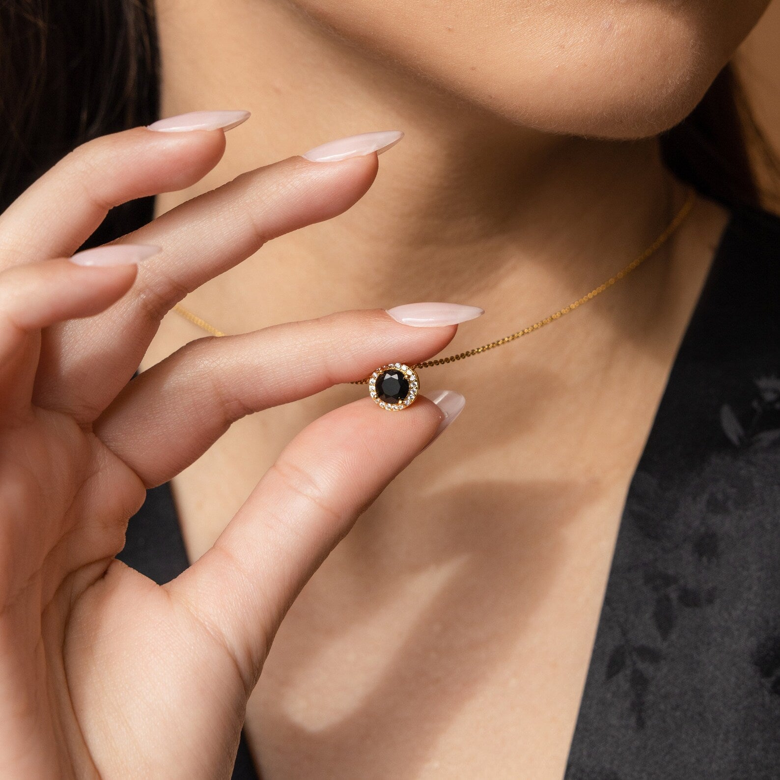 A woman holds the Halo Birthstone Necklace, showcasing a round black gemstone pendant encircled by shimmering diamonds.