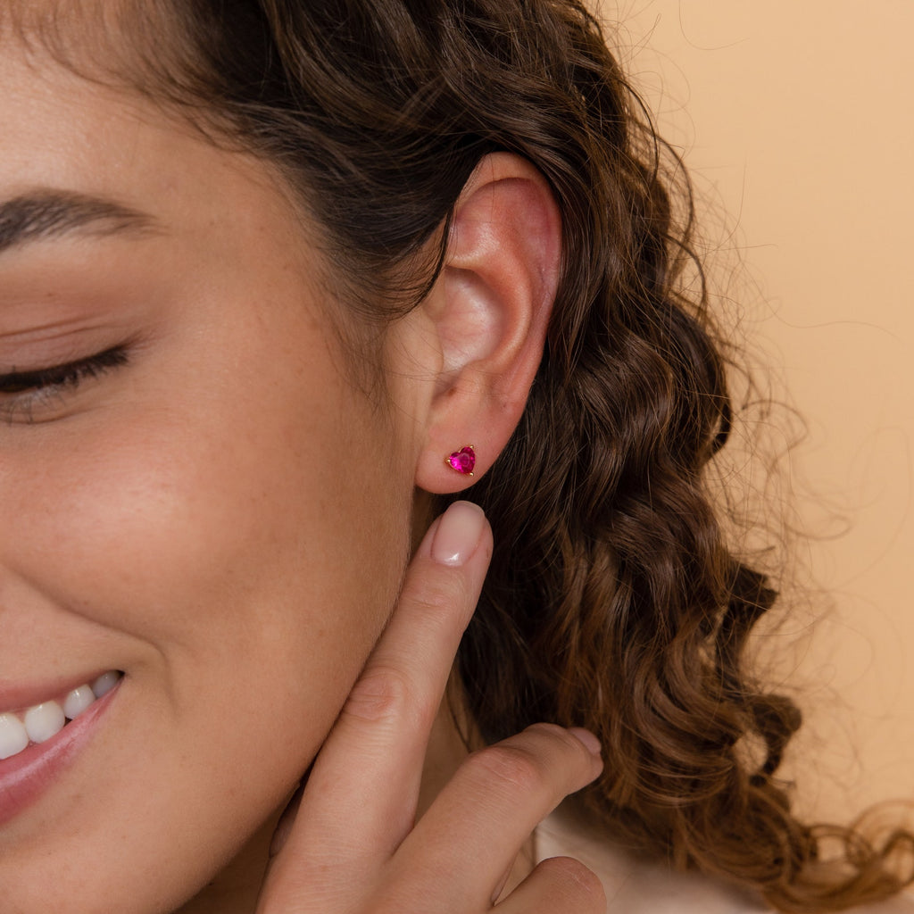 A woman smiles, touching her Ruby Heart Studs—a pair of small pink earrings—with her curly hair visible, creating a perfect look for date night.